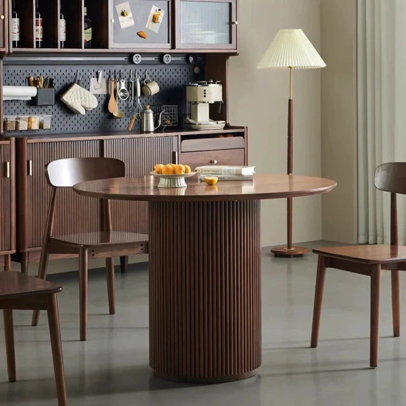 A Round Wood Pedestal Dining Table in a Modern Dining Area in Rich Walnut Color, with Served Snacks and books on the table , and Three Solid Wood Dining Chairs